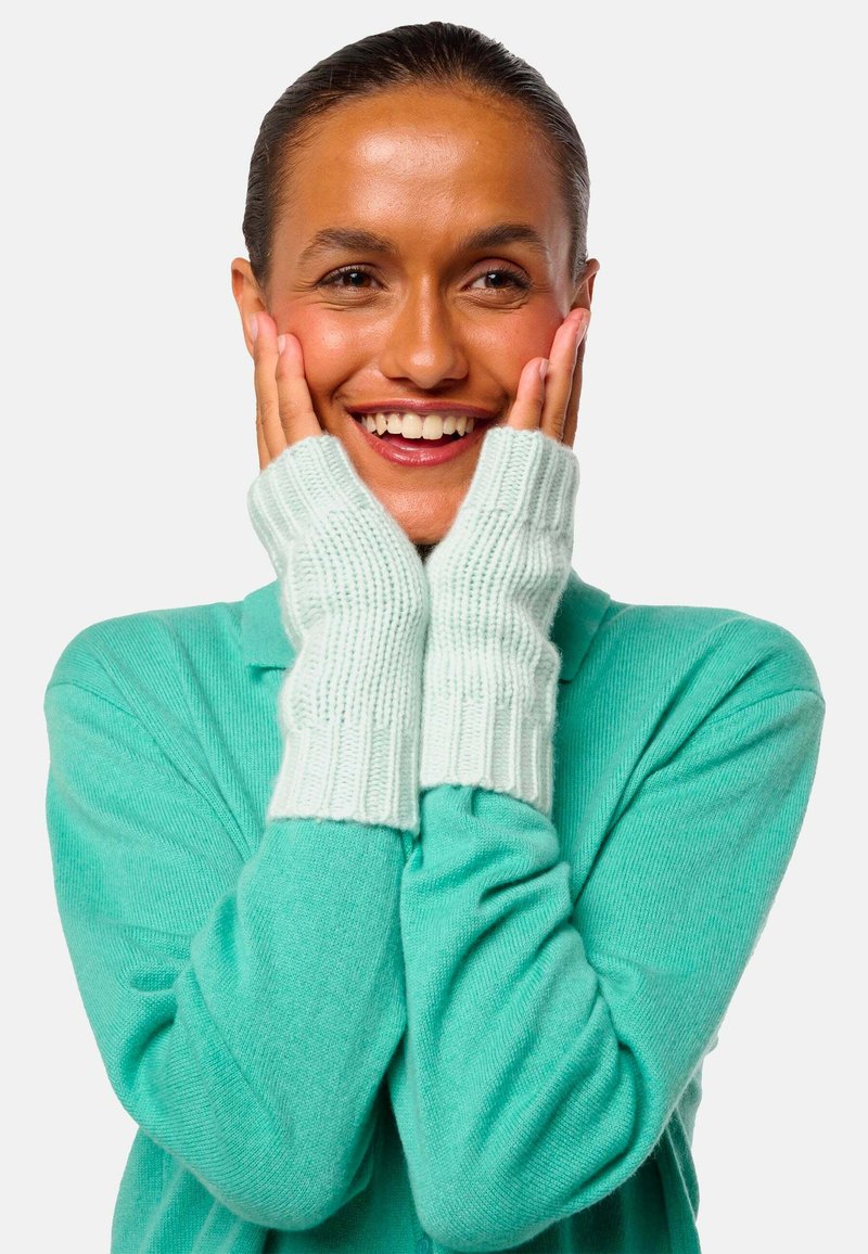 Gants sans doigts bleu clair en tricot doux avec texture côtelée, portés par-dessus un pull turquoise, mettant en valeur la main et le visage.