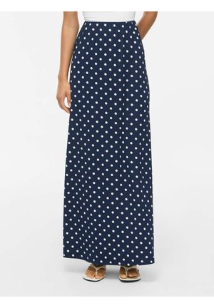 Femme portant une longue jupe bleu marine à pois blancs et des sandales blanches, debout sur un fond blanc uni.