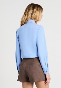 Himmelblaues Langarmhemd mit Kragen, glatter Textur und geknöpften Manschetten, kombiniert mit braunen, gestreiften Shorts mit rückseitigen Taschen.