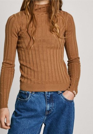 Femme portant un pull marron côtelé à manches longues et un jean bleu en denim, une main dans une poche, sur un fond uni.