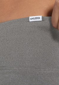 Detailní záběr na ruku upravující šedou žebrovanou tkaninu s bílou etiketou SMILODOX na okraji.