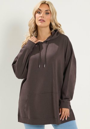 Kvinde med langt blond hår iført en oversized mørkebrun hættetrøje med lomme og snøre, stående mod en ensfarvet baggrund.