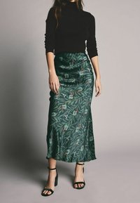 Jupe en satin vert avec motif floral, comprenant des feuilles et des accents rouges. Associée à un pull noir ajusté et des sandales noires à talons carrés.