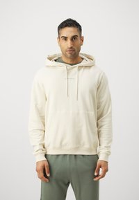 Cream hoodie with a kangaroo pocket, drawstring hood, and subtle branding. Paired with green sweatpants, featuring a relaxed fit.