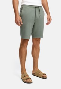 Mand iført olivenfarvede drawstring shorts og sandfarvede ruskinds sandaler, stående mod en ensfarvet hvid baggrund.