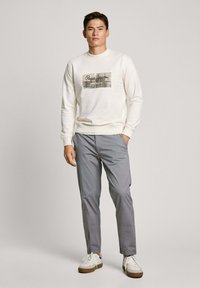 Sudadera blanca con un gráfico impreso en beige y negro, combinada con pantalones grises y zapatillas blancas con suelas marrones.