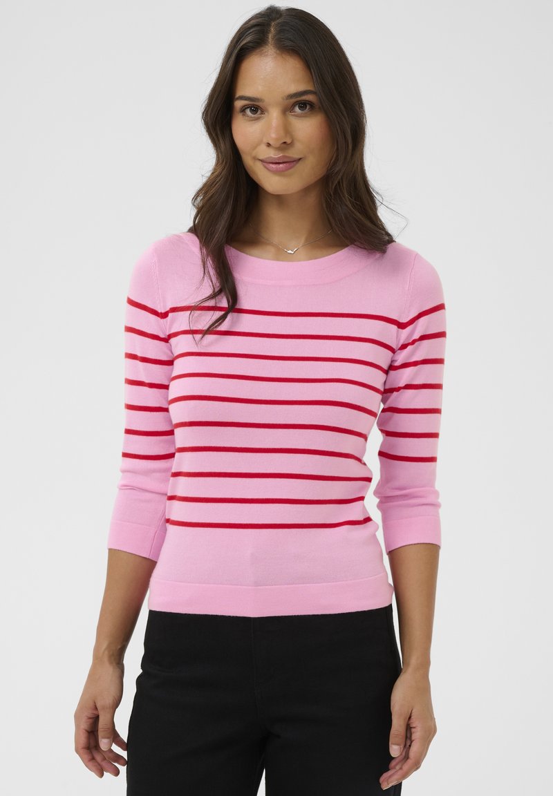 Femme aux longs cheveux foncés portant un pull rose à rayures horizontales rouges et un pantalon noir, debout devant un fond clair uni.