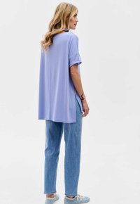 Fioletowa, oversize koszulka z podwiniętymi rękawami, bocznymi rozcięciami oraz prostym krojem, zestawiona z jasnoniebieskimi dżinsami i casualowymi butami.