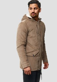 Mand iført en beige corduroy-jakke med hætte og sorte bukser, kigger til sin venstre siden imod en neutral grå baggrund.