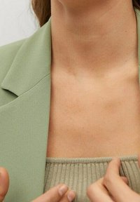 Vaaleanvihreä blazer, jossa on terävät läpät, kerroksittain ribattu beige toppi. Näkyvää ihonväriä kaula-aukon alla, sileä rakenne koko ajan.