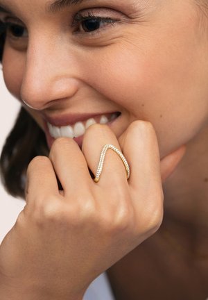 Anillo de oro en forma curva de V, adornado con una fila de pequeñas gemas claras. Llevado en una mano que muestra la textura natural de la piel.