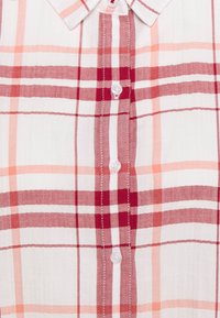 Chemise à boutons blanche avec un motif à carreaux rouges et pêches. Elle dispose d'un col classique et de boutons, fabriquée dans un tissu léger avec une texture lisse.