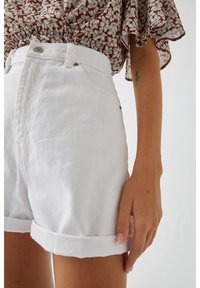 Personne portant un short en denim blanc taille haute et un chemisier marron à fleurs, main détendue le long du corps, tatouage visible sur l'avant-bras.