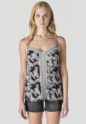 Mujer con cabello rubio largo, vistiendo una blusa sin mangas con estampado de mariposas en blanco y negro y shorts de mezclilla negros, sobre un fondo liso.