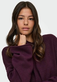 Suéter morado de gran tamaño con una textura suave y puños enrollados, lucido en una pose casual. El cabello está suelto con sutiles ondas.