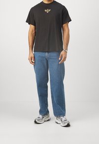 Czarna bawełniana koszulka z złotą grafiką, w zestawieniu z jasnoniebieskimi spodniami o prostym kroju i szarymi trampkami z czarnymi akcentami.