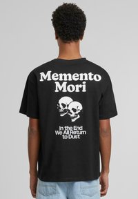 Sorte bomulls-t-skjorte med grafisk design med teksten "Memento Mori", to hodeskaller, og setningen "Til slutt vender vi alle tilbake til støv."