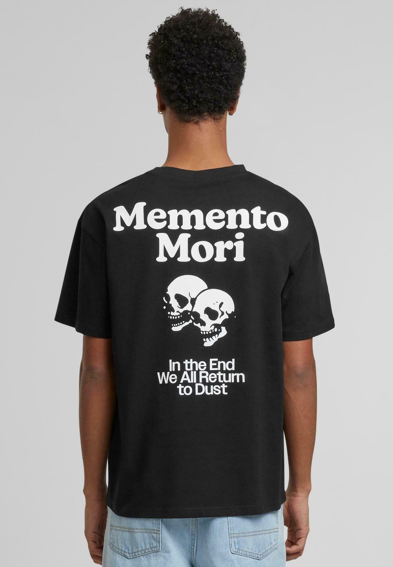 Sorte bomulls-t-skjorte med grafisk design med teksten "Memento Mori", to hodeskaller, og setningen "Til slutt vender vi alle tilbake til støv."