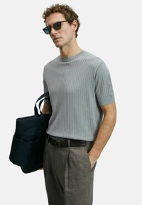 Lichtgrijze ribbel gebreide sweater met korte mouwen, gecombineerd met beige textuurbroek. Het model houdt een marineblauwe tas vast en draagt zwarte zonnebril.