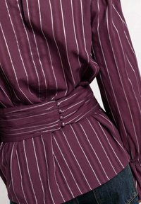 Blusa burdeos con rayas blancas verticales, con una cintura peplum, cinturón de tela con tres botones y mangas largas. Textura suave.
