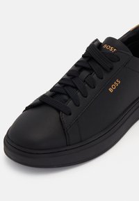 Baskets basses en cuir noir avec lacets noirs, semelle noire et logo "BOSS" doré sur le côté et la languette.