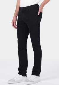 Homme portant un jean noir slim et des baskets blanches, se tenant avec une main dans la poche devant un fond clair.