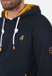 Marineblå hettegenser med gulfôr i hetten, hvite snorer og en brodert gul "S"-logo på brystet. Har en kengurulomme.