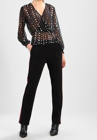 Donna che indossa una blusa nera trasparente con pois, pantaloni neri su misura con strisce rosse laterali e stivaletti neri alla caviglia con dettagli in argento.