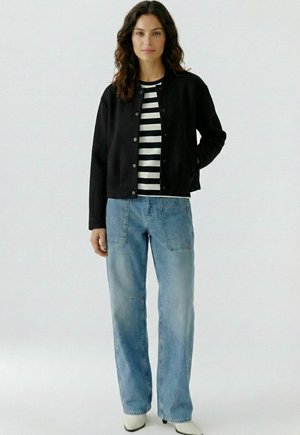 Frau steht, trägt eine schwarze, geknöpfte Jacke, ein schwarz-weiß gestreiftes Hemd, hellblaue Jeans und weiße spitze Stiefel vor einem schlichten Hintergrund.