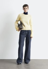 Gelber Strickpullover, darunter ein dunkelblauer Rollkragenpullover, locker sitzende Jeans, hält eine gemusterte dunkle Tasche mit kurzem Griff.