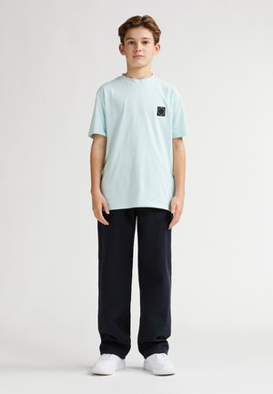 T-shirt di cotone azzurro chiaro con un piccolo logo nero sul petto, abbinata a pantaloni navy a gamba larga e sneakers bianche.