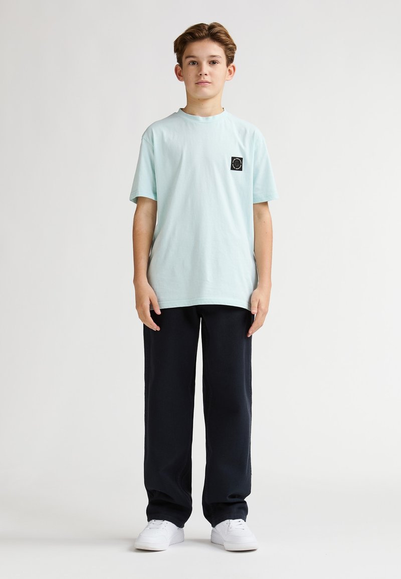 T-shirt di cotone azzurro chiaro con un piccolo logo nero sul petto, abbinata a pantaloni navy a gamba larga e sneakers bianche.