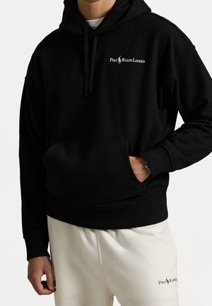Homme portant un sweat à capuche noir Polo Ralph Lauren avec une poche avant et un pantalon de survêtement blanc Polo Ralph Lauren.