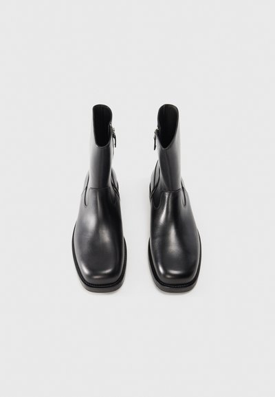 Las botas de tobillo de cuero negro presentan un acabado liso, punta redondeada, cremallera lateral vertical y suela plana con un sutil detalle de costura.