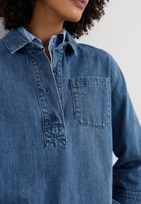 Chemise en denim avec une patte de boutonnage et quatre boutons, dotée d'une poche et d'un col rayé bleu visible en dessous. Couleur bleu foncé et tissu texturé.