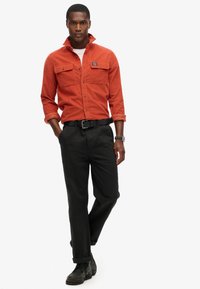 Oranje corduroy overhemd met twee borstzakken, gecombineerd met zwarte broek en zwarte schoenen. Model staat met een neutrale uitdrukking.