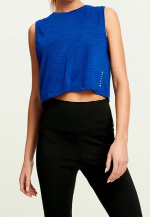 Nainen, joka käyttää sinistä hihattomaa lyhyttä topia, jossa on hienovaraisia aaltoilevia kuvioita, sekä mustia korkeavyötäröisiä leggingssejä, seisoo yksinkertaisen vaalean taustan edessä.