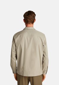 Camicia button-up beige chiaro con maniche lunghe, colletto standard e una texture liscia. Presenta bottoni sui polsini e una cucitura a yoke sul retro.