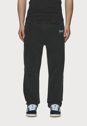 Mand iført sorte jeans med vide ben med "Pescado"-logo på højre lomme og sorte sneakers med hvide snørebånd og såler.