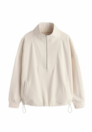 Kermaankeltainen pusero, jossa on korkea kaulus, puolipitkä vetoketju edessä, sivutaskut ja säädettävä kiristysnauha helmassa. Sileä pinta ja rento istuvuus.