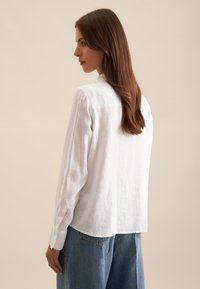 Chemise blanche en lin à manches longues avec col, poignets boutonnés et coupe décontractée, assortie à un pantalon en denim bleu ample.