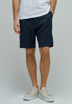 Hombre con pantalones cortos de denim oscuro, zapatillas blancas y camiseta blanca, de pie con una mano en el bolsillo frente a un fondo liso.