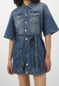 Denim shirt dress in dark blue with short sleeves, button front, two chest pockets, and a tie belt at the waist. Slightly frayed hem.