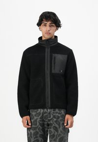 Tânăr îmbrăcat cu o jachetă de fleece neagră cu fermoar frontal și buzunar pe piept, purtând pantaloni gri cu carouri digitale, stând pe un fundal alb.