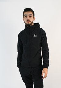 Chaqueta ligera negra con cierre de cremallera, capucha ajustable y puños elásticos. Incluye un logo blanco en el pecho. Textura suave.
