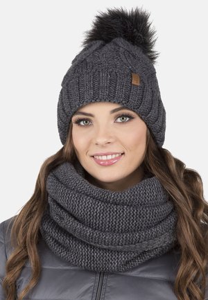 Cappello in maglia grigio con pom pom in pelliccia sintetica sopra, abbinato a una sciarpa infinita in maglia grigio spessa, entrambi con modelli tessurizzati.