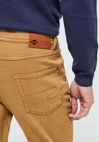 Pantalons beiges en denim avec une étiquette en cuir à la taille. Couture des poches visible et un design légèrement arrondi sur la poche arrière.