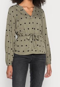Blouse à manches longues olive avec des pois noirs, décolleté en V, taille cintrée avec un lien, portée avec un jean gris foncé.