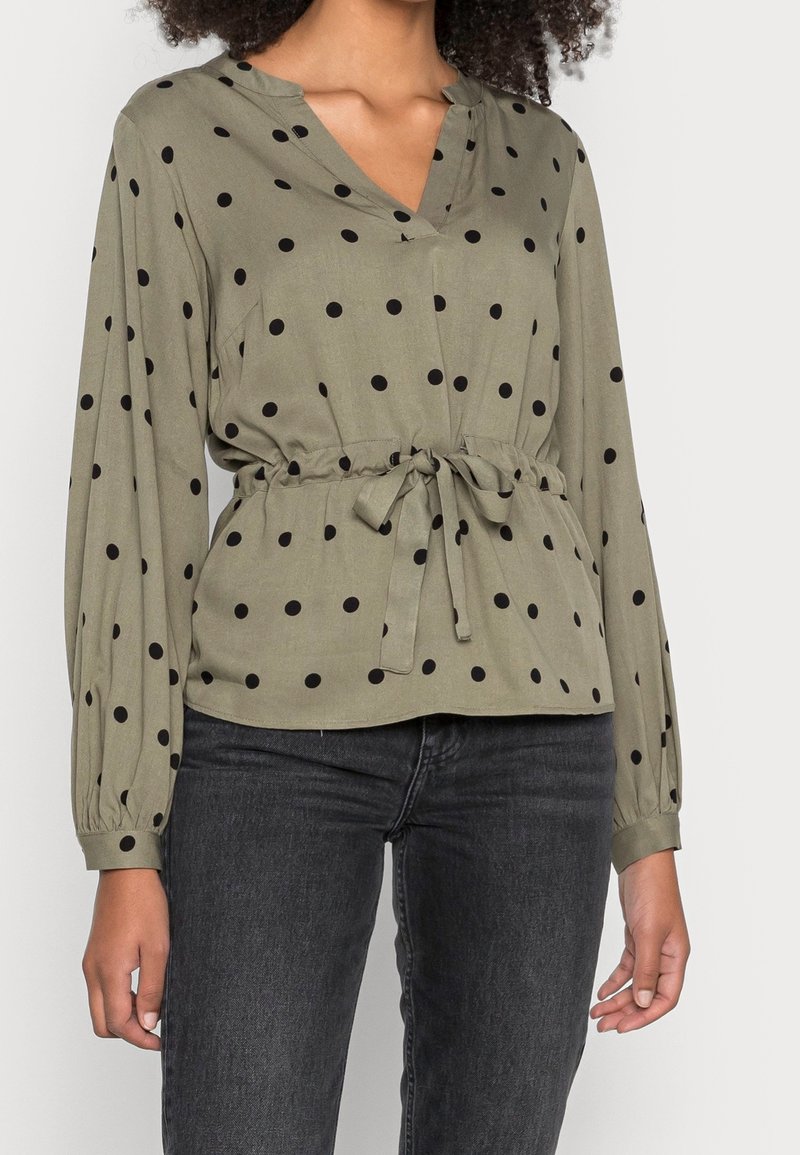 Blouse à manches longues olive avec des pois noirs, décolleté en V, taille cintrée avec un lien, portée avec un jean gris foncé.