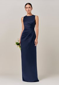 Večerna obleka iz mornarsko modrega satena s prilegajočo se silhueto, brez rokavov in stranjo z nagubanim detajlom. Model drži šopek belih cvetov.